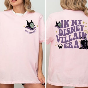 May include: Pink t-shirt featuring a purple and green image of Maleficent from Disney's Sleeping Beauty. The back of the shirt displays the text "In My Disney Villain Era" in purple with a gold crown and a purple and black image of Maleficent.
