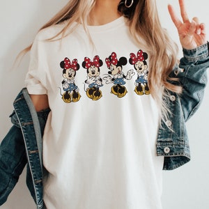 May include: White t-shirt with a graphic of four Minnie Mouse characters wearing red bows and blue skirts. The characters are arranged in a row and are facing forward.