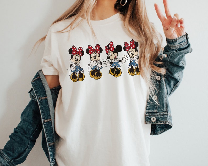 May include: White t-shirt with a graphic of four Minnie Mouse characters wearing red bows and blue skirts. The characters are arranged in a row and are facing forward.
