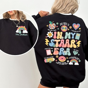 May include: Black sweatshirt with a colorful graphic print that says "In My Staar Era". The graphic includes a rainbow, a donut, a pencil, a diamond, a book, a flower, and a heart. The text "Mrs. Jacobson" is printed on the front of the sweatshirt.