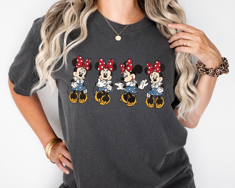 May include: A dark gray t-shirt with a graphic of four Mickey Mouse characters wearing red bows and blue dresses. The characters are in a row, facing forward, and are smiling.