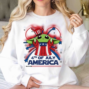 Puede incluir: Sudadera blanca con un personaje extraterrestre verde que lleva un sombrero de T&iacute;o Sam rojo, blanco y azul y sostiene un fuego artificial. El texto "4th of July America" est&aacute; impreso en la camiseta.