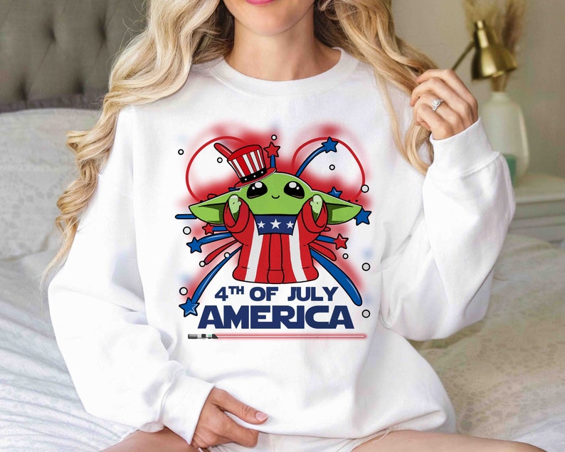 Puede incluir: Sudadera blanca con un personaje extraterrestre verde que lleva un sombrero de T&iacute;o Sam rojo, blanco y azul y sostiene un fuego artificial. El texto "4th of July America" est&aacute; impreso en la camiseta.