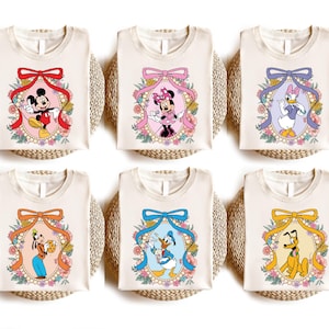 May include: Six white t-shirts with different Disney character designs. Each shirt features a character in a floral frame with a bow at the top. The characters include Mickey Mouse, Minnie Mouse, Donald Duck, Daisy Duck, Goofy, and Pluto.