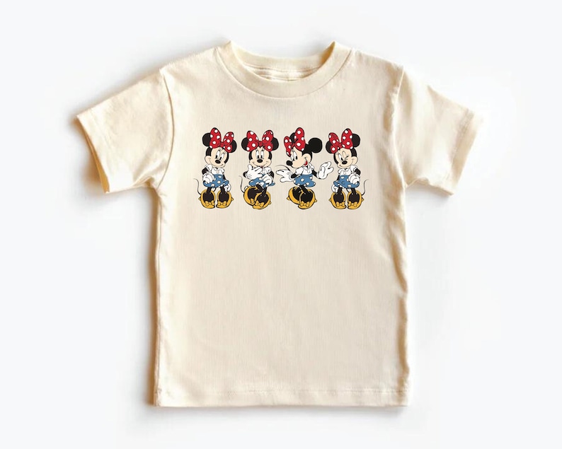 May include: A cream colored t-shirt with a graphic of four Mickey Mouse characters wearing red and white polka dot bows. The characters are all facing forward and have their arms outstretched.