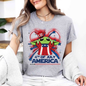 Puede incluir: Una camiseta gris con una imagen de dibujos animados de un extraterrestre verde que lleva un sombrero rojo, blanco y azul y sostiene un fuego artificial. El texto "4th of July America" est&aacute; impreso debajo del extraterrestre.