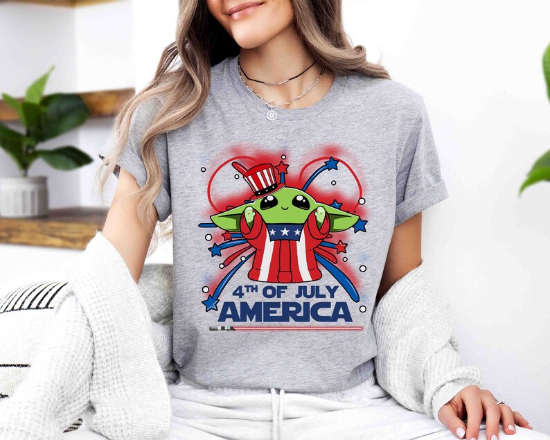 Puede incluir: Una camiseta gris con una imagen de dibujos animados de un extraterrestre verde que lleva un sombrero rojo, blanco y azul y sostiene un fuego artificial. El texto "4th of July America" est&aacute; impreso debajo del extraterrestre.