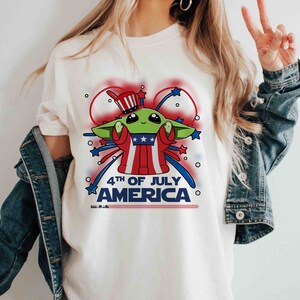 Puede incluir: Camiseta blanca con una imagen de dibujos animados de un extraterrestre verde con un sombrero de t&iacute;o Sam rojo, blanco y azul y sosteniendo un fuego artificial. El texto "4th of July America" est&aacute; impreso debajo de la imagen.