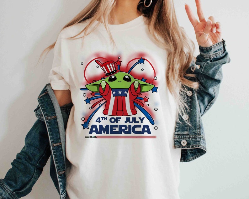 Puede incluir: Camiseta blanca con una imagen de dibujos animados de un extraterrestre verde con un sombrero de t&iacute;o Sam rojo, blanco y azul y sosteniendo un fuego artificial. El texto "4th of July America" est&aacute; impreso debajo de la imagen.