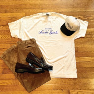 Puede incluir: Un atuendo informal que presenta una camiseta blanca con "Directed by David Lynch" en escritura azul, pantalones de pana marrones y mocasines de cuero marrón oscuro. También se incluye una gorra de béisbol blanca y negra con un logotipo.