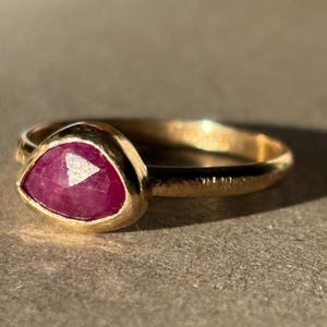 May include: A gold ring with a pink ruby gemstone set in a bezel setting. The ring has a textured band.