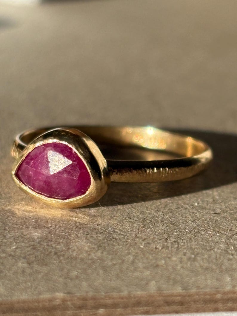 May include: A gold ring with a pink ruby gemstone set in a bezel setting. The ring has a textured band.