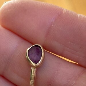May include: A gold ring with a purple gemstone set in a bezel setting. The ring band is textured.