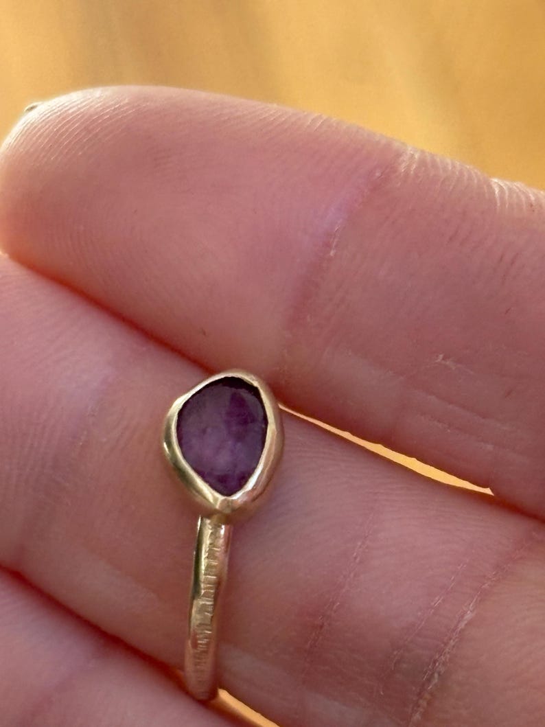 May include: A gold ring with a purple gemstone set in a bezel setting. The ring band is textured.