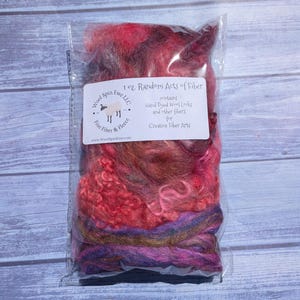 May include: A clear plastic bag containing 1 oz. of hand-dyed wool locks and fibers in shades of pink, red, purple, and brown. A white label on the front reads: 1 oz. Random Acts of Fiber, contains Hand Dyed Wool Locks and other fibers for Creative Fiber Arts, Wool Spin Ewe LLC.