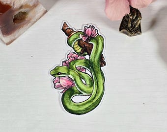 Pode incluir: Um autocolante com uma cobra verde enrolada num galho castanho, adornada com flores cor-de-rosa. A cobra tem um aspeto detalhado, semelhante a uma aguarela. O autocolante está numa superfície branca.
