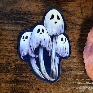 May include: A sticker featuring a cluster of ghost mushrooms. The mushrooms have white caps with black eyes and mouths, and long, flowing, white veils. The stems are a dark blue-grey color. The sticker has a dark blue border.
