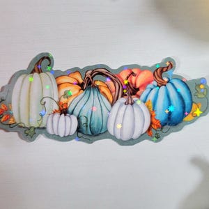 May include: A sticker featuring a row of five pumpkins in various shades of blue, white, and orange. The pumpkins are decorated with glitter and have a fall theme.