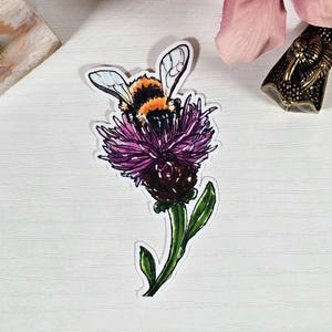 May include: A sticker featuring a detailed illustration of a bumblebee on a purple thistle flower. The bee has black and orange stripes, with white and blue wings. The flower has a dark brown center and green stem.
