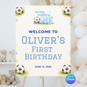 以下が含まれることがあります： 四隅にサッカーボールのイラストが描かれた長方形の誕生日用サインボード。青い文字でOur little rookie is ONE, WELCOME TO OLIVER'S FIRST BIRTHDAY、日付JUNE 15, 2026と記されており、背景は白いテクスチャ加工が施されています。