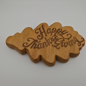 May include: A wooden Thanksgiving decoration in the shape of a leaf. The words "Happy Thanksgiving" are carved into the wood in a decorative font. The wood has a warm, natural tone.