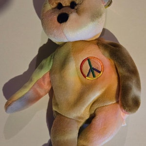 May include: A tie-dye plush bear with a peace sign on its chest. The bear is brown, yellow, pink, and green. The peace sign is green, yellow, red, and blue.