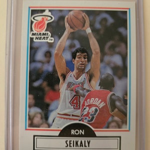 May include: A basketball trading card featuring Ron Seikaly, a center for the Miami Heat. The card is from the 1990 Fleer basketball card set. Seikaly is wearing a red and white Miami Heat jersey with the number 4 on the front. He is jumping up to shoot a basketball. The card has a blue border and a white background.