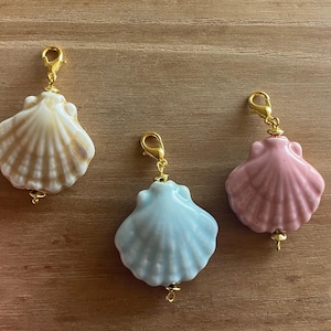 May include: Three seashell charms in cream, light blue, and pink, each with a gold-colored clasp and decorative bead. The charms are arranged on a wooden surface, showcasing their delicate details and colors.