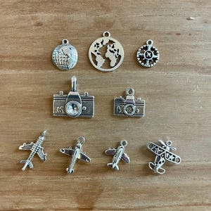 May include: A set of silver charm pendants featuring travel-themed designs, including a globe, compass, camera, airplane, and a small plane.