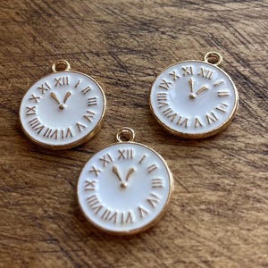 May include: Three gold-tone clock charms with white enamel faces and black Roman numeral markings. The charms have a loop at the top for attaching to a necklace or bracelet.