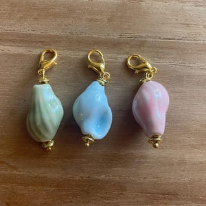 May include: Three seashell charms in pastel colors: light green, light blue, and pink. Each charm has a gold-colored clasp and decorative accents. The charms are displayed on a wooden surface.