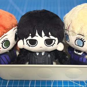 May include: Three plush dolls in a small, light-coloured container. The dolls have large eyes and are dressed in dark blue and black outfits. One doll has orange hair, one has black hair, and one has blonde hair.