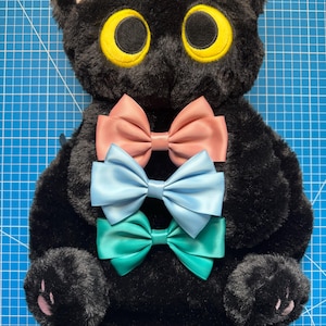Puede incluir: Bolso negro con forma de gato de peluche con grandes ojos amarillos, orejas internas rosas y tres lazos de satén en rosa, azul claro y verde azulado. El bolso tiene una correa de cadena y detalles en forma de pata.
