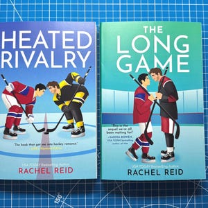 May include: Two books with hockey-themed covers. "Heated Rivalry" shows two hockey players facing off, while "The Long Game" depicts two players holding hockey sticks. Both books are by Rachel Reid, a USA Today Bestselling Author.