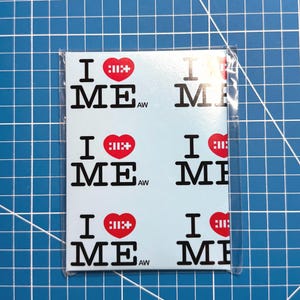 May include: A light blue card with the text "I ME" in black, repeated multiple times. The heart symbol is red with a white cross design. The card is in a clear plastic sleeve.
