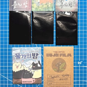 Puede incluir: Colección de tarjetas fotográficas y mini soportes con texto e ilustraciones coreanas. Las tarjetas tienen embalaje negro. Una tarjeta tiene dibujos animados de gatos. Otra tiene una ilustración botánica. El fondo es una cuadrícula azul.