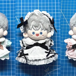 May include: Three plush dolls with gray hair and blue eyes, each wearing a different outfit. The left doll wears a light blue dress with a brown polka-dot bow. The center doll wears a black and white checkered dress with a matching bonnet. The right doll wears a pink dress with a black polka-dot bow and the word Angel embroidered on the front.