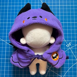 May include: A plush doll wearing a purple hooded cape with black horns and orange accents. The cape features a Halloween-themed emblem on the side. The doll is on a blue grid surface.