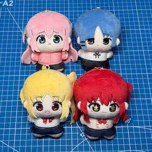 May include: Four plush character keychains with large heads and small bodies. One has pink hair, one has blue hair, one has yellow hair, and one has red hair. Each figure wears a white top and a dark skirt, featuring embroidered facial details.