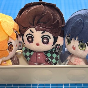 May include: Three plush dolls with large eyes and detailed facial features sit in a small, light-coloured container. The dolls have yellow, brown, and blue hair. The dolls are in a container on a blue surface.