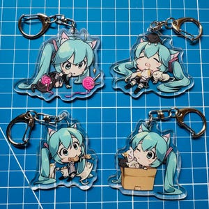 May include: Four clear acrylic keychains featuring a cartoon character with turquoise hair and cat ears. Each keychain has a silver clasp. The character is depicted with various items, including yarn, cats, and food.
