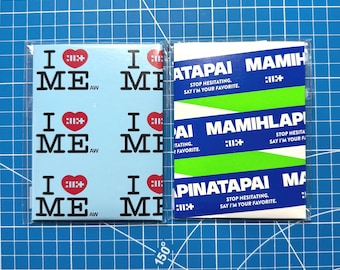 May include: Two rectangular stickers in clear plastic sleeves. The left sticker is light blue with a repeating pattern of black text reading I HEART ME and a red heart. The right sticker features horizontal blue and lime green stripes with white text reading MAMIHLAPINATAPAI and STOP HESITATING. SAY I'M YOUR FAVORITE.