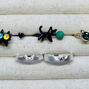 May include: Five cat-themed rings, including two silver rings with cat faces, a black ring with a cat holding a ball of yarn, a black ring with a cat face, and a gold ring with a green gemstone and cat ears.