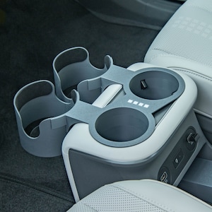May include: A gray car cup holder with two cup holders and a charging port. The cup holder is made of plastic and has a textured surface.