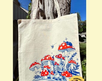 Puede incluir: Una bolsa de lona blanca con una ilustración colorida de setas e insectos. La bolsa tiene un diseño azul y rojo con el texto "Sourbunny Studios" en la esquina inferior derecha.