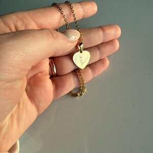 May include: A gold-colored necklace featuring a heart-shaped pendant. The pendant has a delicate floral engraving. The necklace is held in a hand, showcasing the chain and the pendant's design. The chain is a delicate link style.