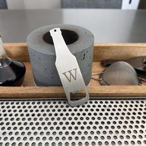 May include: A silver bottle opener shaped like a beer bottle with the letter "W" engraved on the front. The opener is sitting on a wooden tray with a metal grate.