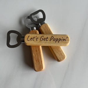 May include: Three wooden bottle openers with metal bottle opener tops. The middle bottle opener has the text "Let's Get Poppin'" engraved on the wooden handle.