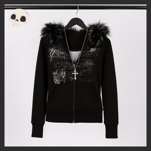 May include: Black zip-up hoodie with faux fur trim on the hood. The hoodie features a graphic design on the front and a silver cross pendant necklace. The hoodie is hanging on a black hanger.