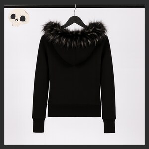 May include: Black hooded sweatshirt with a faux fur trim around the hood. The sweatshirt is hanging on a black hanger against a white background. The cuffs and hem are ribbed. A cartoon skull is in the upper left corner.